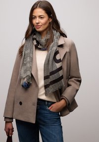 Sort-hvidt mønstret tørklæde med blå accenter, lagt over en beige frakke. Tørklædet har geometriske mønstre og en blød tekstur.