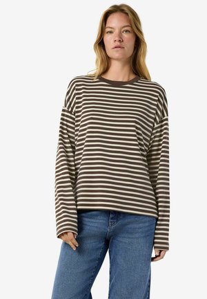 Vrouw met blond haar die een bruin-wit horizontaal gestreept shirt met lange mouwen en blauwe spijkerbroek draagt, staand tegen een effen achtergrond.