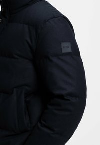 Giubbotto puffer blu marina con cuciture orizzontali, chiusura frontale con zip e automatici, tasca laterale e un'etichetta logata BOSS in tessuto sulla manica sinistra.