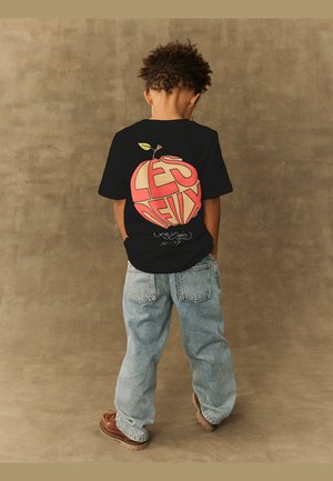 T-shirt noir avec un graphique coloré de pomme et du texte "LES DEUX". Porté avec un jean en denim bleu clair et des chaussures marron, vu de dos.