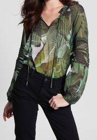 Nainen, jolla on päällään läpinäkyvä vihreä paita, jossa on trooppisia lehtikuvioita, ja mustat housut, käsi lantiolla.