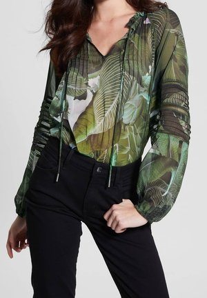 Kvinde iført en gennemsigtig grøn bluse med tropiske bladmønstre og sorte bukser, hånd hviler på hoften.