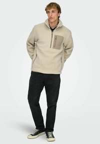 Beige värinen fleece-pusero, jossa korkea kaulus, neljännesvetoketju ja kontrastivärinen tasku. Yhdistetty mustiin farkkuihin ja mustiin lenkkikengiin.