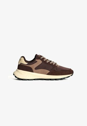 La zapatilla de correr de ante marrón y malla presenta una suela crema, paneles en contraste y cordones. El diseño incluye un cuello acolchado y una suela texturizada.