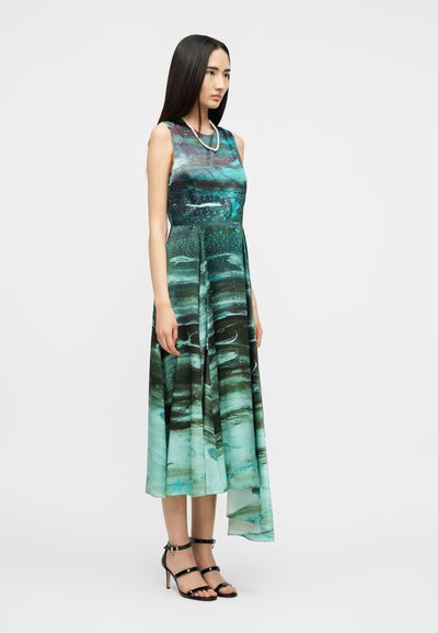 Γυναίκα στέκεται φορώντας ένα αμάνικο μεσοφόρι σε τιρκουάζ και μαύρο με αφηρημένο σχέδιο, με ασύμμετρο τελείωμα και μαύρα πέδιλα με λουράκια.