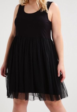 Robe noire sans manches avec corsage ajusté et jupe en tulle superposée portée par une personne debout sur un fond uni.