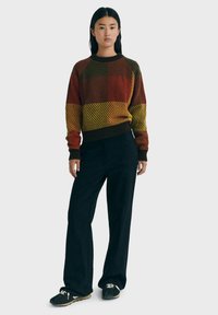 Maglione lavorato a maglia con motivo a quadri multicolore in rosso, arancione, giallo e verde. Abbinato a pantaloni larghi neri e scarpe nere.
