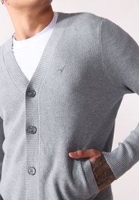 Světle šedý cardigan s ribovanou texturou, V-výstřihem, pěti knoflíky a bočními kapsami. Obsahuje decentní výšivku loga na hrudi.