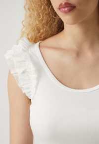 Hvitt rynket tank top med volanghvitt stoff på venstre skulder. Glatt tekstur med rund hals og tilpasset design.