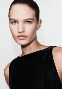 Młoda osoba z wygładzonymi ciemnymi włosami, nosząca bezrękawnik z czarnego aksamitu, patrzy bezpośrednio w kamerę na tle jednolitego tła.