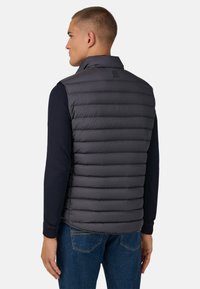 Gilet grigio imbottito con colletto alto e design trapuntato orizzontale. Indossato sopra una camicia a maniche lunghe scura, con pantaloni in denim blu.