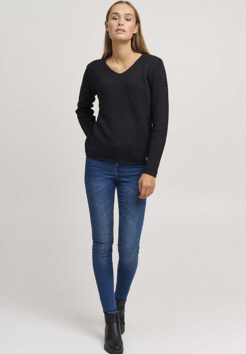 Zwarte V-hals trui met een textuurpatroon, lange mouwen en geribbelde zoom, gecombineerd met blauwe skinny jeans en zwarte enkellaarsjes.