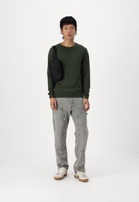 Grønn strikkegenser, grå baggy jeans med synlige sømmer, hvite joggesko med sideakenter, og en svart crossbody-veske.