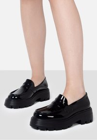 Mocasines negros de charol con suela gruesa, que presentan un acabado suave, acentos estriados y un diseño sin cordones.