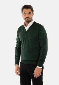 Maglione verde a scollo a V in un punto a nido d'ape, taglio aderente, abbinato a una camicia bianca con colletto. Pantaloni neri completano l'outfit.