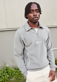 Γκρίζο φλις πουλόβερ με κλείσιμο με φερμουάρ στον λαιμό, με λευκό λογότυπο "BARÇA" και σήμα Nike, επισημασμένο από πλαινές τσέπες με φερμουάρ.