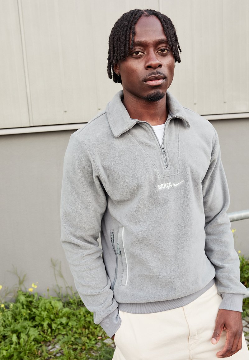 Γκρίζο φλις πουλόβερ με κλείσιμο με φερμουάρ στον λαιμό, με λευκό λογότυπο "BARÇA" και σήμα Nike, επισημασμένο από πλαινές τσέπες με φερμουάρ.