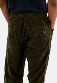 Pantaloni in velluto a coste verde oliva con una superficie testurizzata, vita elastica e due grandi tasche posteriori. Presenta un piccolo logo.