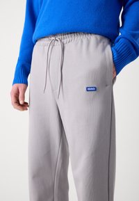 Pantalones deportivos grises con cintura ajustable que presentan un pequeño logo azul de "HUGO". La textura parece suave y lisa, combinando con un suéter azul.