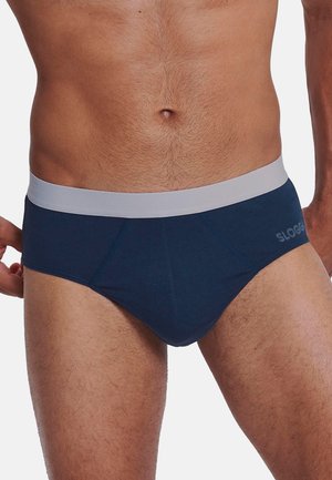 Torso maschile indossando slip blu navy con una cintura in vita grigio chiaro, che mostra gli avambracci parzialmente e la parte superiore delle cosce su uno sfondo semplice.
