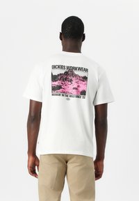 Camiseta blanca de algodón con un estampado gráfico de montañas en negro y rosa en la parte posterior, con el texto "DICKIES WORKWEAR" arriba.