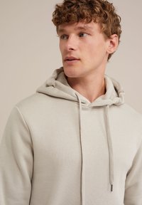 Svetlo siva kapuca iz soft čvrste tkanine, sa prednjim džepom, podesivim vezicama i opuštenim krojem sa rebrastim manžetama.