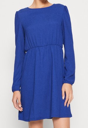 Femme portant une robe texturée bleu roi, longueur genou, manches longues, avec une taille froncée et un encolure ronde.