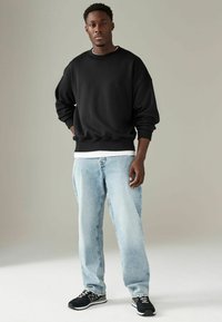 Zwarte crewneck sweatshirt met een losse pasvorm, blauwe spijkerbroek en zwarte sneakers met witte accenten. Model staat tegen een neutrale achtergrond.