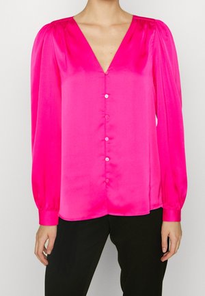 Kirkkaan pinkki satiinipusero, jossa pitkät puffihihat, napitettava etuosa ja syvä V-pääntie, yhdistetty mustiin housuihin.