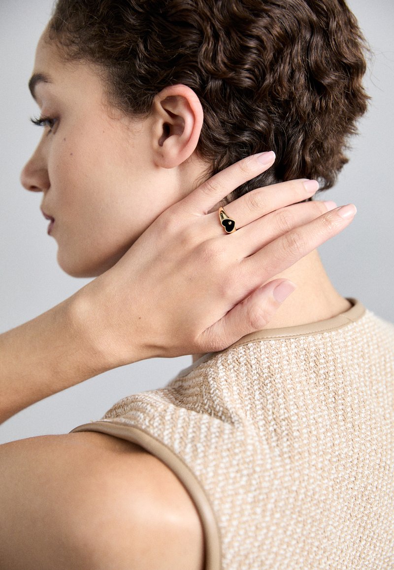 Zlatý prsteň s čiernym srdcovým drahokamom, nosený na ruke, ktorá sa dotýka krku. Texturovaný béžový pletený top s hladkým hnedým okrajom.