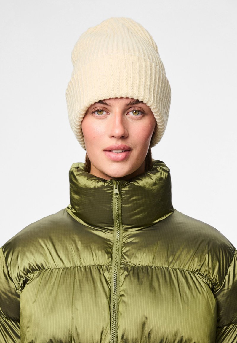 Zelená, lesklá vatovaná bunda s vysokým golierom a zipsom, doplnená o mäkkú krémovú rebrovanú čiapku. Model s svetlými očami a neutrálnym pozadím.