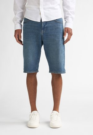 Man in halflange blauwe spijkershorts, wit overhemd met knopen en witte sneakers, staand tegen een effen witte achtergrond.