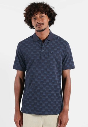Homme aux cheveux bouclés portant un polo bleu foncé à motifs et un pantalon clair, debout devant un fond blanc.