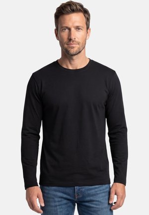 Man met kort haar en lichte baard, gekleed in een effen zwarte trui met lange mouwen en blauwe spijkerbroek, kijkt recht vooruit tegen een neutrale achtergrond.