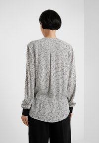 Blouse fleurie à base blanche, motifs minuscules noirs et marron, manches longues, détail froncé au dos, et poignets noirs.