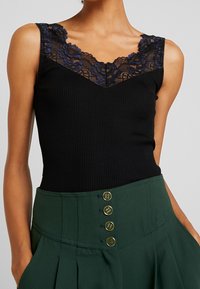 Czarny prążkowany top na ramiączkach z głębokim dekoltem w kształcie litery V ozdobiony niebieską koronką w kwiaty. Połączony z zielonymi spodniami z wysokim stanem i złotymi guzikami.