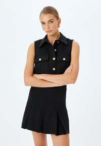 Schwarzes ärmelloses Kleid aus strukturiertem Material, mit goldenen Knöpfen, zwei Brusttaschen und einem Faltenrockdesign.