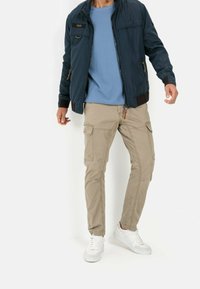 Mężczyzna w granatowej kurtce, jasnoniebieskiej koszuli, beżowych spodniach cargo i białych trampkach, stojący na białym tle.