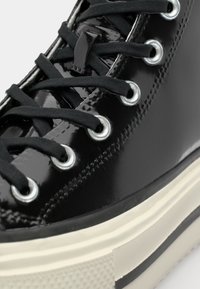Nærbilde av svarte lakk lær sneakers med svarte snører, sølvnitter, en teksturert gummisåle og en kontrasterende beige stripe.