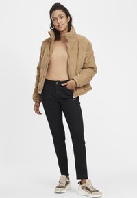 Barna kordbársony bombázódzseki, bordázott bézs pulóver, fekete farmer és világosbarna cipők hálós részletekkel. A modell semleges háttér előtt áll.