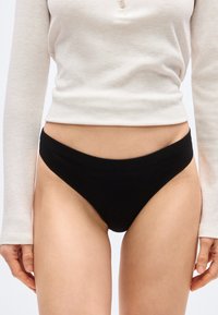 Γυναίκα που φοράει μαύρο εσώρουχο με ψηλό κόψιμο και μακρυμάνικο ανοιχτό μπεζ ριμπ πλεκτό μπλουζάκι με λεπτομέρεια κουμπιών, εμφανίζεται από τη μέση έως το μέσο του κορμού.