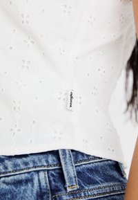 Bijela pamučna bluza s izvezenim rupicama i cvjetnim uzorcima, vidljiv oznaka s natpisom "Wrangler", uparena s plavim trapericama.