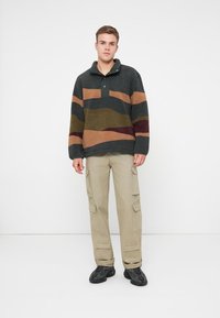 Fleece-Pullover mit Kragen, ausgestattet mit dunkelgrünen und beige horizontalen Streifen, kombiniert mit beige Cargo-Hosen und schwarzen Sneakers.