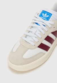 Adidas tenisice s bež antilopom i gornjim dijelom od bijele kože. Imaju crvene horizontalne pruge, bijele vezice i plavu oznaku na jeziku.