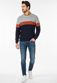 Suéter en gris, azul marino y óxido con rayas horizontales, textura de punto, cuello redondo y detalle de logo. Combinado con jeans azules y zapatillas blancas.