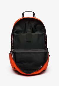 Pomarańczowy plecak z czarnym wnętrzem, zamykany na zamek błyskawiczny, z kieszeniami wewnętrznymi; ma siatkową sekcję oraz górny uchwyt do noszenia.