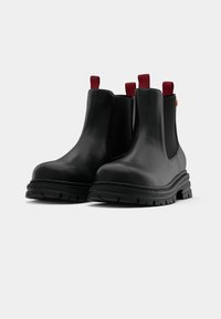 Botas Chelsea de goma negra con acabado texturizado, que cuentan con paneles laterales elásticos, pestañas rojas y una suela de goma negra gruesa.