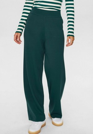 Dunkelgrüne, weit geschnittene Hose mit einem elastischen Bund, aus weichem Stoff, kombiniert mit weißen Sneakers und einem gestreiften Oberteil.