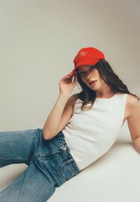 Czerwona czapka baseballowa z haftowanym napisem, zestawiona z białą bluzką bez rękawów oraz niebieskimi dżinsami z wysokim stanem. Gładkie materiały, swobodny styl.