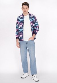 Jachetă bomber multicolor cu imprimeu de palmier în nuanțe de albastru, roz și negru. Purtați-o peste un tricou alb, cu blugi deschiși la culoare și adidași albi.